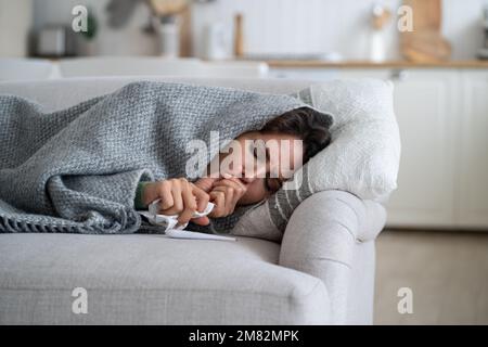 Kranke erschöpfte Frau, die auf der Couch liegt, in eine Decke gewickelt und hustet, nachdem sie infiziert wurde gefährliche Grippe Stockfoto