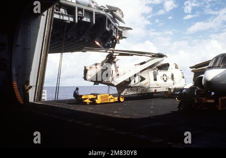 Ein Sattelschlepper SD-1D wird verwendet, um einen SH-3H Sea King von Helicopter Anti-U-Boot Squadron 8 in einem abgesenkten Aufzug an Bord des Flugzeugträgers USS CONSTELLATION (CV 64) zu manövrieren. Auf der rechten Seite befindet sich ein Wikingerflugzeug S-3A. Der Spediteur nimmt an Flottenübung 85 Teil. Basis: USS Constellation (CV 64) Land: Pazifik (POC) Stockfoto