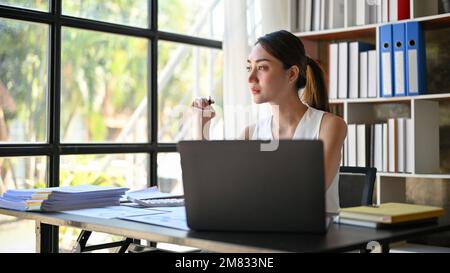 Professionelle und seriöse asiatische Geschäftsfrau, die nachdenklich ist, aus dem Fenster blickt und an ihrem Schreibtisch beharrlich über ihr Unternehmensprojekt nachdenkt. Stockfoto