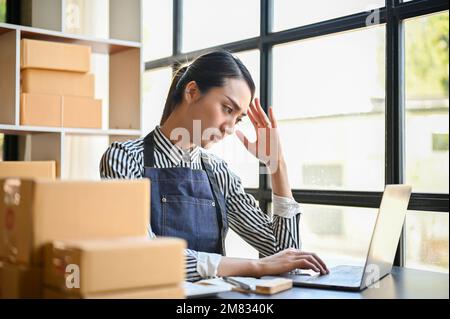Gestresste und konzentrierte asiatische Millennials-E-Commerce-Unternehmerin oder Online-Shop-Besitzerin, die ihren Laptop benutzt und ihre Online-Bestellung überprüft Stockfoto