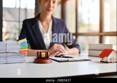 Selektives Fokusbild, Richterhammer, Vertrag, Dokumente, Hausmodell und Bürobedarf auf dem Tisch über verschwommenem Hintergrund einer Anwältin. Stockfoto