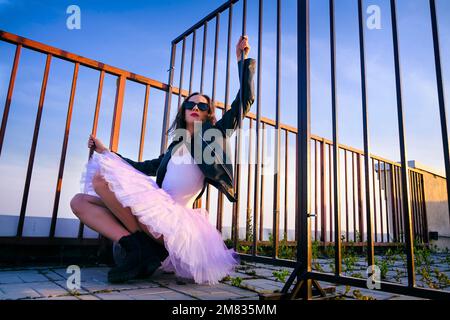 Eine Ballerina in Tutu, die bei Sonnenuntergang am Zaun auf dem Dach sitzt, in Stiefeln und einer Jacke Stockfoto