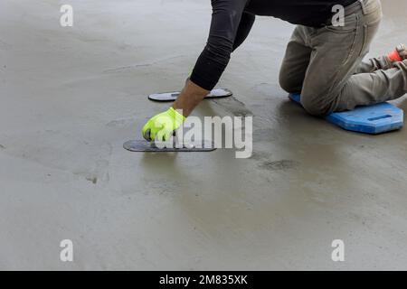 Während der Maurer eine Stahlkelle in der Hand hält, glättet er während der Arbeit Putzbeton auf Zementboden Stockfoto