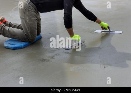 Während er eine Maurerkelle aus Stahl in seiner Hand hält, glättet der Maurer den Putzbeton auf den Zementboden Stockfoto