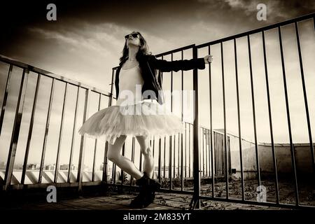 Eine Ballerina in Tutu steht am Zaun auf dem Dach bei Sonnenuntergang in Stiefeln und einer Jacke Stockfoto