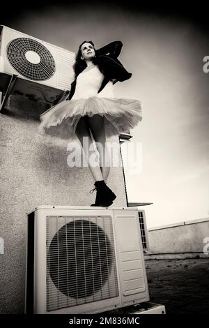 Eine Ballerina in Tutu, Jacke und Stiefeln kletterte auf die Klimaanlage und posierte gegen den Himmel Stockfoto