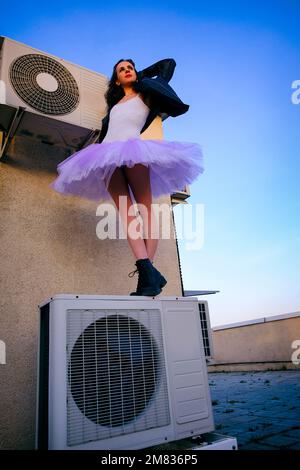 Eine Ballerina in Tutu, Jacke und Stiefeln kletterte auf die Klimaanlage und posierte gegen den Himmel Stockfoto