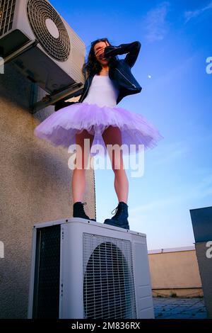 Eine Ballerina in Tutu, Jacke und Stiefeln kletterte auf die Klimaanlage und posierte gegen den Himmel Stockfoto