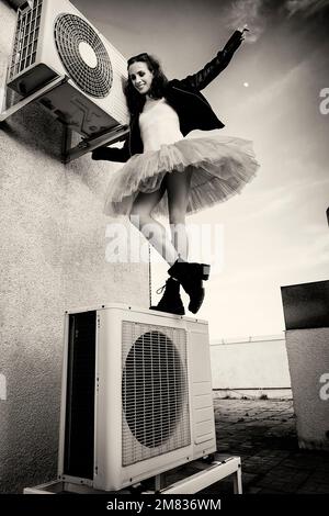 Eine Ballerina in Tutu, Jacke und Stiefeln kletterte auf die Klimaanlage und posierte gegen den Himmel Stockfoto