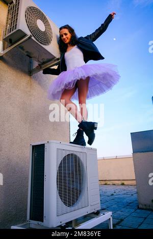 Eine Ballerina in Tutu, Jacke und Stiefeln kletterte auf die Klimaanlage und posierte gegen den Himmel Stockfoto