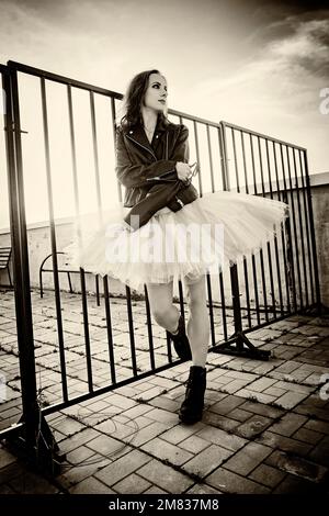 Eine Ballerina in Tutu steht am Zaun auf dem Dach bei Sonnenuntergang in Stiefeln und einer Jacke Stockfoto