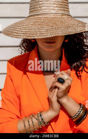 Stilvolle Frau in einem orangefarbenen Anzug und einem Strohhut. An den Händen sind Schmuckringe und Armbänder auf einem weiß gestreiften Hintergrund. Stockfoto