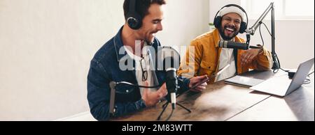 Fröhliche Podcaster, die lachen und sich in einem Heimstudio amüsieren. Zwei glückliche junge Männer, die eine Live-Audioübertragung moderieren. Zwei junge Ersteller von Inhalten Stockfoto