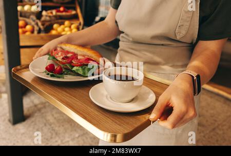 Unbekannte junge Kellnerin, die ein Tablett mit einem Sandwich und einer Tasse Kaffee trug. Junge Frau, die als Kellnerin in einem Fast-Food-Restaurant arbeitet. Stockfoto