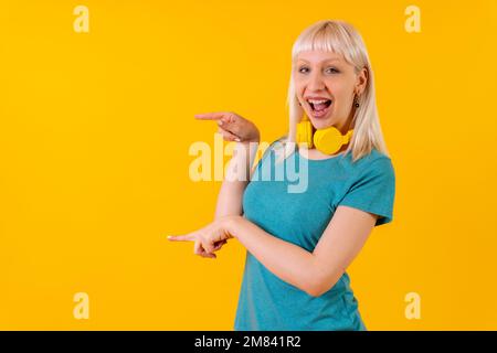 Eine blonde Weiße mit Kopfhörern am Hals, die auf den gelben Hintergrund zeigen Stockfoto