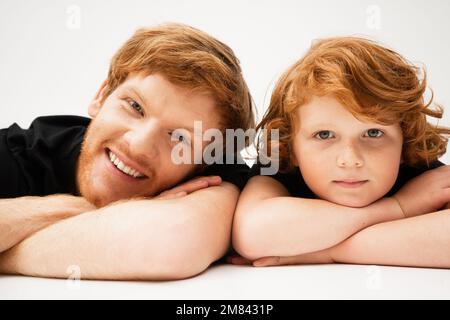 Fröhlicher junger Mann mit rothaarigem Sohn, der mit gekreuzten Armen auf hellgrauem Hintergrund liegt, Stockbild Stockfoto