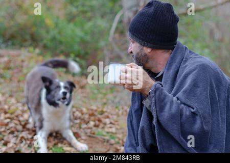 Ein bärtiger Mann mit einer Decke, der eine Tasse Kaffee trinkt, mit seinem Hund im Hintergrund Stockfoto