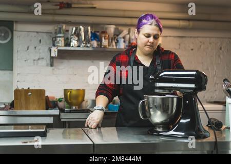 Bäckerin mit lila Haaren, die Produkte in einer Schüssel in der Küche mischt, Bäckerei, Kochkonzept. Hochwertiges Foto Stockfoto
