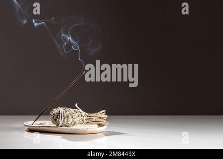 Brennender aromatischer Incense Smoky Stick und ein Bündel Salvia Salvia Salbe auf weißem Tablett auf dunklem minimalistischem Hintergrund. Aromatherapie-Rauch für Meditation und Re Stockfoto