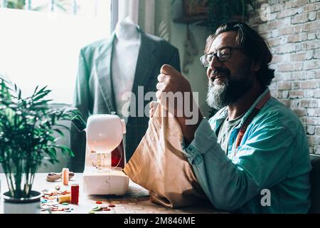 Glücklicher Arbeiter in der Werkstatt. Schneide den Mann allein mit der Nähmaschine und lächle während der Arbeit. Alternative handgefertigte Arbeit. Erwachsene Männer in der Schneiderei Stockfoto