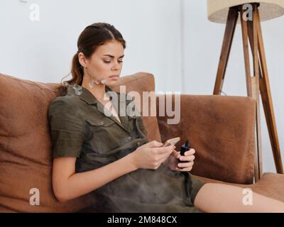 Freiberufliche Bloggerin, die auf dem Sofa sitzt, mit Telefon in der Hand und rauchenden E-Zigarettenvape, stilvolle Inneneinrichtung. Stockfoto