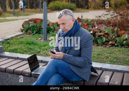 Ein alter, grauhaariger Geschäftsmann mit Brille sitzt im Herbst im Stadtpark auf einer Bank, arbeitet an einem Laptop und unterhält sich am Telefon. Stockfoto