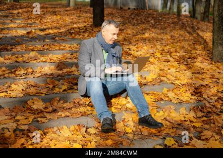 Porträt eines alten Geschäftsmannes, der auf einer Treppe sitzt, die mit gelben Blättern im Wald bedeckt ist, und auf dem Laptop tippt. Stockfoto