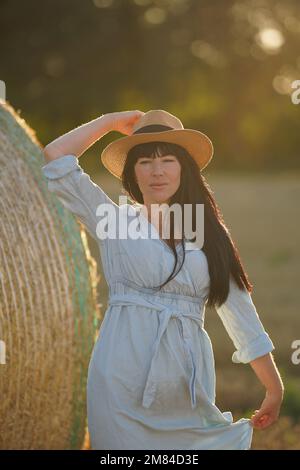 Ein ukrainisches Mädchen in Nationalkleidung mit schwarzem dickem Haar und einem leichten Kleid hält eine Hand auf einem Strohhut auf ihrem Kopf Stockfoto