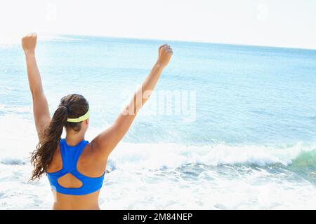 Ich hab's geschafft. Rückblick auf eine junge Frau, die auf das Meer blickt, mit erhobenen Armen zur Feier. Stockfoto