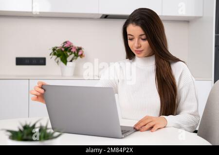 Eine junge, schöne Frau sitzt an ihrem Arbeitsplatz und öffnet das Notebook, um online zu arbeiten. Freiberufliche Arbeit zu Hause. Stockfoto