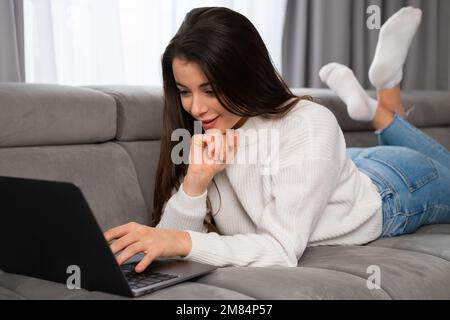 Glückliche junge Frau, die zu Hause auf dem Sofa liegt und auf dem Laptop-Bildschirm sitzt, Nachrichten liest, Videos ansieht, mit Freunden chattet Stockfoto