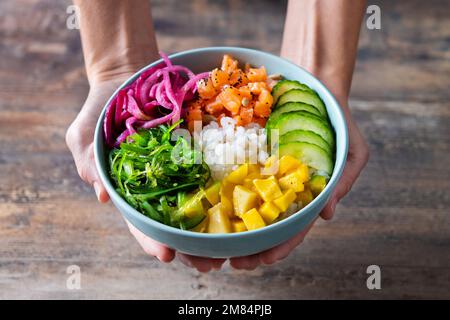 Frauenhände halten eine Schüssel mit Reis, Lachs, Gurke, Mango, Zwiebeln, Wachsalat, Mohnsamen und Sonnenblumenkernen auf Holzhintergrund Stockfoto