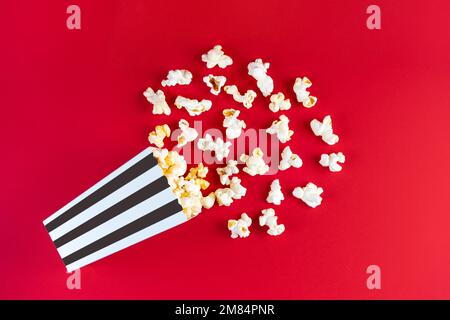 Leckeres Käsepopcorn, das aus einem schwarz gestreiften Eimer fällt, isoliert auf rotem Hintergrund. Streuung von Popcornkörnern. Filme, Kino und Mitmachen Stockfoto