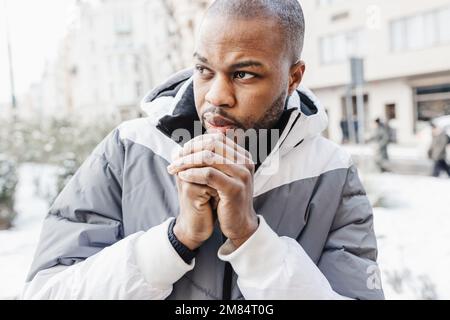 Junger Afrikaner in legerer weißer und blauer Kleidung, wärmt seine kalten Hände mit Atem. Straßenporträt mit Schnee in der Wintersaison. Lifestyle, Stadtleben Konzept. Schließen Stockfoto