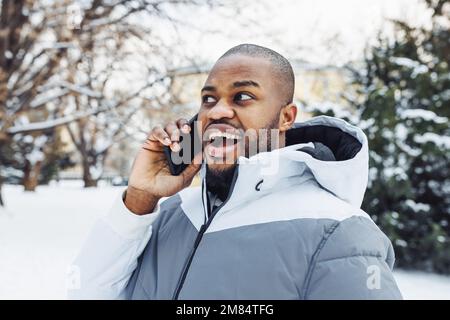 Glücklicher, überraschter afroamerikanischer Mann in legerer weißer Sportkleidung, der am Telefon telefoniert, lächelt und im Park oder auf der Straße draußen schockiert ist. Wintersaison mit Schnee Stockfoto