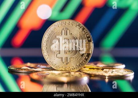 Goldene Bitmünze, die auf einer Spiegeloberfläche steht und einen verschwommenen Hintergrund reflektiert, mit Kryptografik mit grünen und roten Kerzenleuchtern. Konzept des Marktes für den Handel mit Kryptowährungen Stockfoto