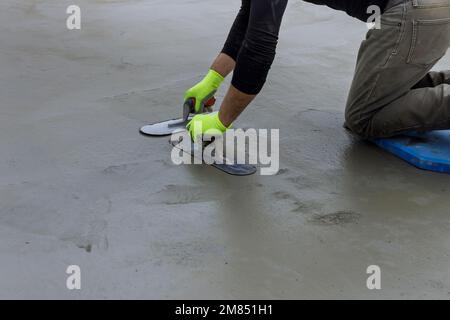 Während er eine Stahlkelle in den Händen hält, glättet der Maurer verputzten Beton auf dem Zementboden Stockfoto