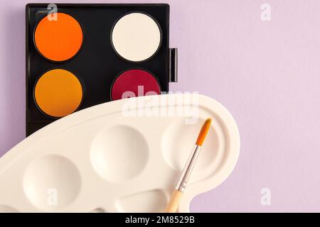 Warme Wasserfarben und Pinsel mit Farbpalette auf violettem Hintergrund. Malerei und Kunst Hobbyausrüstung. Platz für Text kopieren Stockfoto