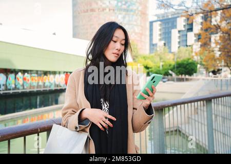 Eine seriöse asiatische Geschäftsfrau, die in der Handy-App eine SMS im Freien sucht. Unglückliches chinesisches Mädchen, das Nachrichten auf dem Smartphone liest. Hochwertiges Foto Stockfoto