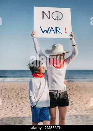 Eine Mutter und ihr asiatischer Sohn sehen sich an, während die Mutter ein Schild mit der Aufschrift "kein Krieg" hochhält. Kein Krieg- und Friedenskonzept. Ihrem Sohn Werte beibringen. ver Stockfoto