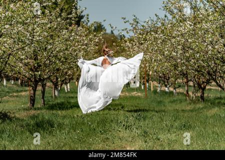 Eine Frau in einem weißen Kleid läuft durch einen blühenden Kirschgarten. Das lange Kleid fliegt zur Seite, die Braut jubelt im Leben. Stockfoto