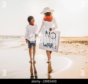 Eine Mutter und ihr asiatischer Sohn schauen sich an und gehen Hand in Hand am Strand bei Sonnenuntergang entlang, mit einem Schild mit der Aufschrift „Nein zum Krieg“. Krieg und Erbse beenden Stockfoto