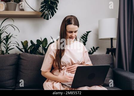 Glückliche, schöne schwangere Frau, die auf dem gemütlichen Sofa sitzt und mit dem Laptop Videogespräche mit der Familie führt, mit Geburtshelfer/Gynäkologe spricht, Empfang Stockfoto