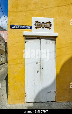 WILLEMSTAD, CURACAO - 22. NOVEMBER 2007: Das Straßenschild Hanchi di Bientu und Conscientiesteeg über der weißen Tür in einem gelben Gebäude in der Allee Stockfoto