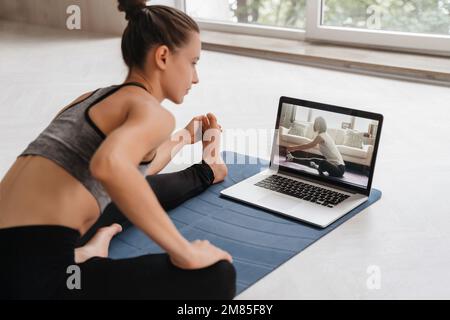 Fit Frau in Sportbekleidung, die zu Hause Yoga auf der Trainingsmatte macht und ein Tutorial für Laptops verwendet. Die Trainerin hat einen virtuellen Yoga-Kurs am Computer. Gesundes und aktives Lifestyle-Konzept. Online-Schulung zu Hause Stockfoto