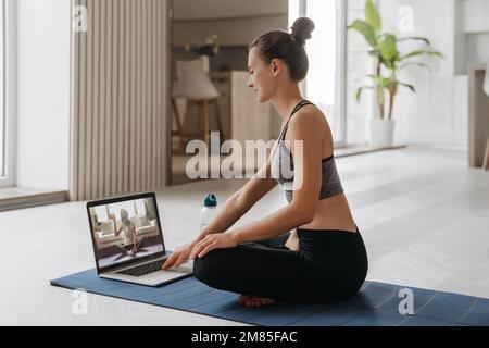 Yoga-Lehrer, der zu Hause einen virtuellen Yoga-Kurs auf einer Videokonferenz durchführt. Wunderschöne Frau, die online Yoga in ihrem Wohnzimmer mit einem Laptop praktiziert. Fitness- und Trainingskonzept. Online-Schulung Stockfoto