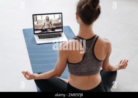 Nahaufnahme einer jungen sportlichen Frau, die online mit einem Laptop zu Hause Yoga praktiziert. Yoga-Lehrer führt Videotraining per Videokonferenz durch. Meditation, Sukhasana, entspann dich. Konzept eines gesunden Lebensstils Stockfoto