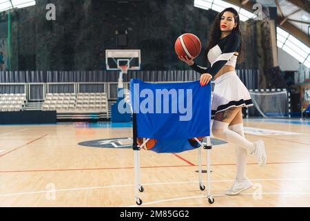 Eine junge, attraktive Cheerleaderin posiert selbstbewusst vor einem blauen Basketballcontainer. Sie hält selbstbewusst einen Basketball in einer Hand und hebt ein Bein, um ihre Stärke und Leichtathletik zu demonstrieren. Stockfoto