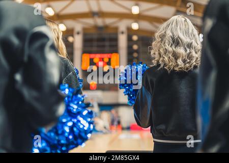 Eine Gruppe Cheerleader von hinten in einem Hallenstadion während eines Spiels. Sie halten ein paar Pom Poms in der Hand. Die Mädchen tragen schwarze Jacken und passgenaue Hosen. Stockfoto