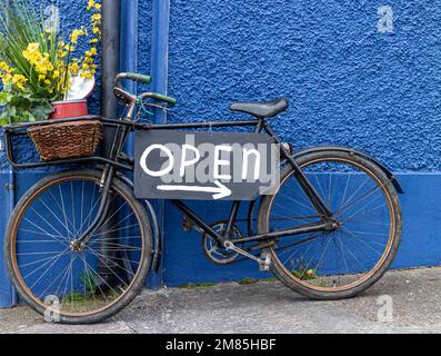 Offenes Schild, öffnendes Schild und Hinweisschild auf einem alten schwarzen Fahrrad, dekoriert mit Blumen und an einer blauen Wand Stockfoto